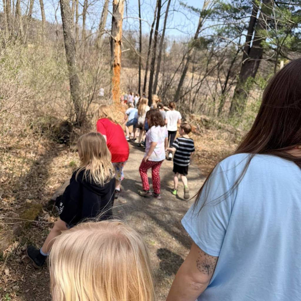 A group of Unity 2nd graders and their teacher walking down a sloped dirt path on the nature trail. The photo is taken from behind the group, showing a line of students in colorful clothing heading toward a sun-drenched wooded area during their Earth Day hike.