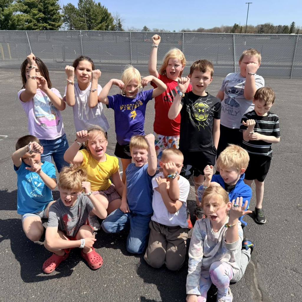 A group of about a dozen Unity 2nd grade students posing together on the school blacktop. Several students are flexing their muscles and smiling confidently for the camera, showing off the paper-bead bracelets they made from recycled book pages for Earth Day.