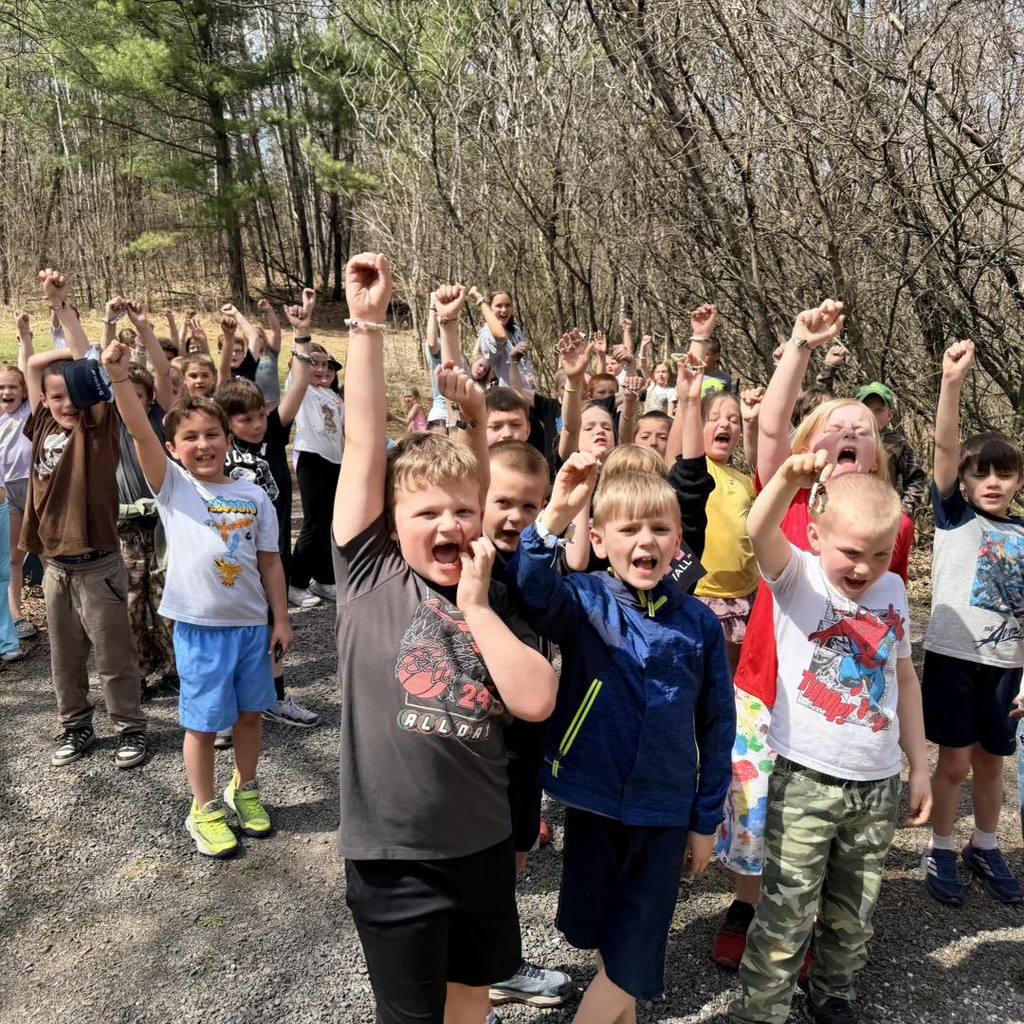 A large group of Unity 2nd grade students cheering on a gravel nature trail. Many of the children have their fists raised in the air in celebration, surrounded by a forest of budding trees during their Earth Day nature hike.