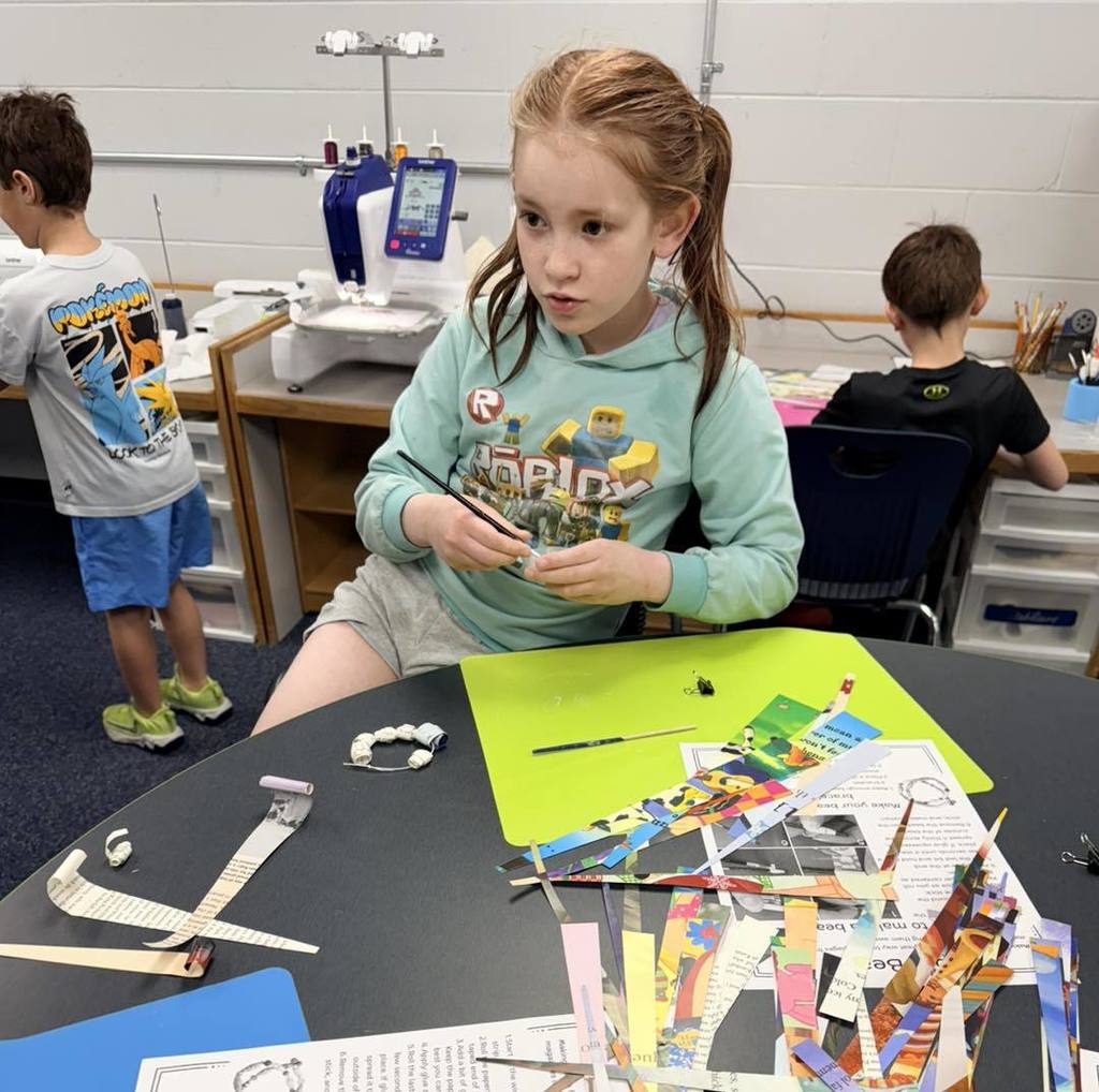 A Unity 2nd grade girl in a teal hoodie sits at a table, carefully rolling a strip of paper from a recycled book to create a bead. Spread out before her are many colorful, triangular paper strips and several completed beads, showcasing the Earth Day craft project in progress.