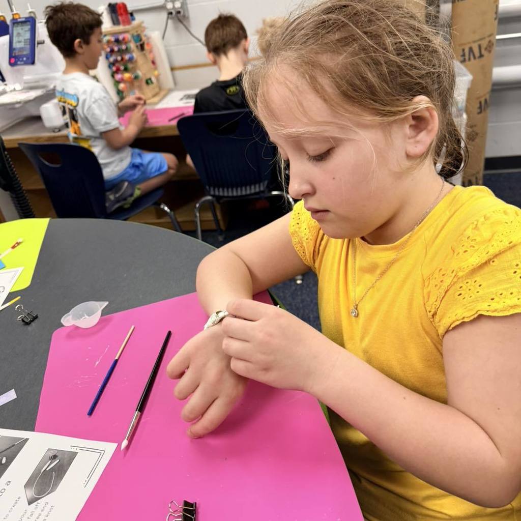 A Unity 2nd grade girl in a yellow shirt sits at a table, focused on sliding a handmade paper bead onto a string to create a bracelet. She is working on a bright pink craft mat with tools and recycled paper instructions nearby, as other students work on their Earth Day projects in the background.