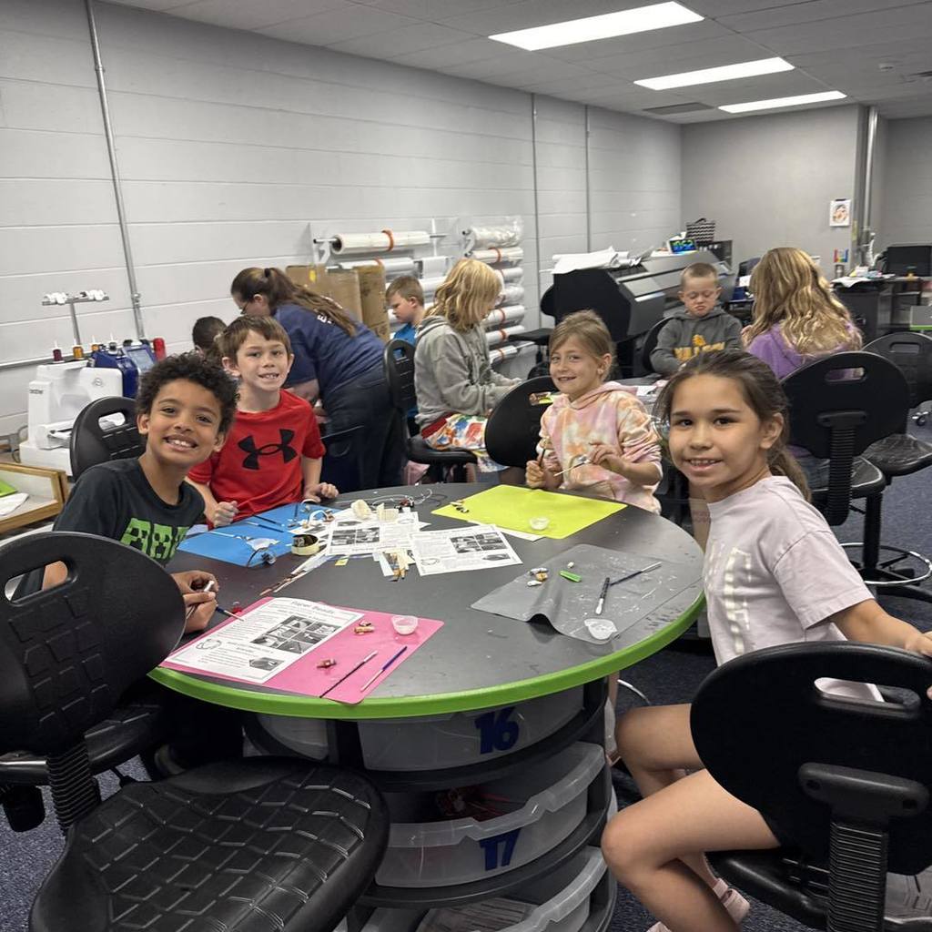 A group of Unity 2nd grade students sitting at a large circular table in a classroom, smiling while making Earth Day bracelets. The table is covered with green craft mats, paper-bead instruction sheets, and colorful strips of recycled book pages. In the background, classroom storage and crafting equipment are visible.