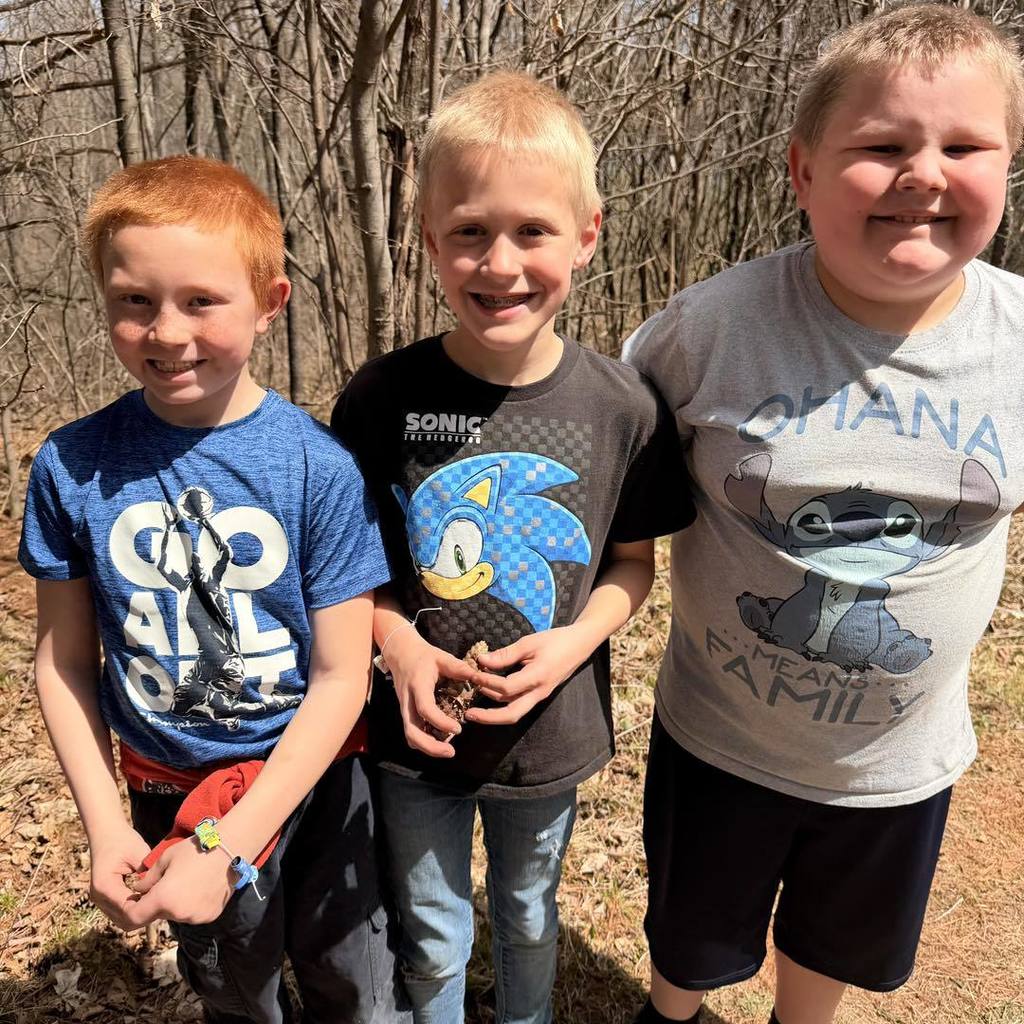 Three Unity 2nd grade boys standing together and smiling on a sunlit nature trail. The boys are wearing graphic t-shirts, including a blue shirt, a black Sonic the Hedgehog shirt, and a grey Stitch shirt. One student is wearing his handmade recycled paper-bead bracelet while they enjoy their Earth Day hike.