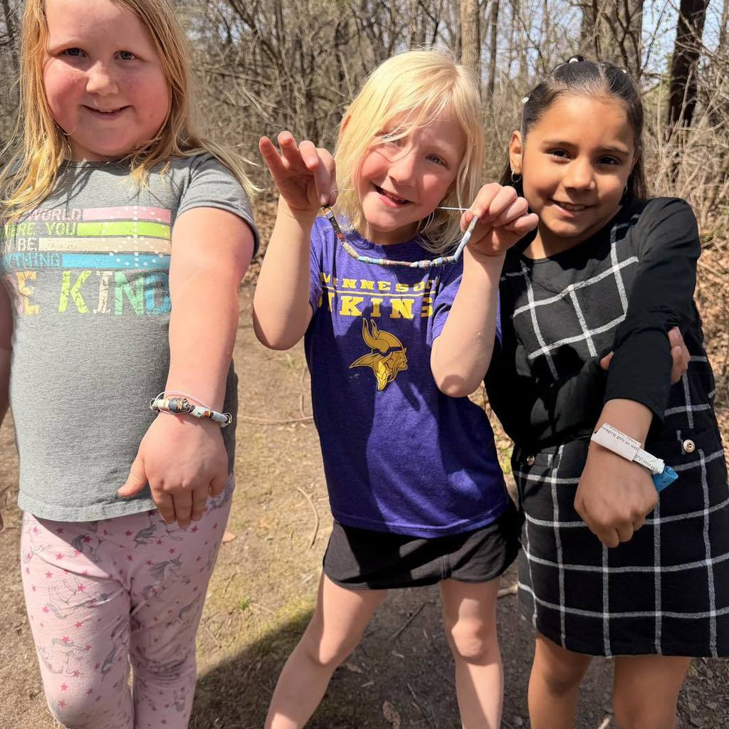 Three Unity 2nd grade girls smiling on the nature trail while displaying their handmade Earth Day bracelets. The girl in the center wears a purple Vikings shirt and holds her bracelet up, while her classmates show theirs on their wrists, highlighting the colorful recycled paper beads they created in class.
