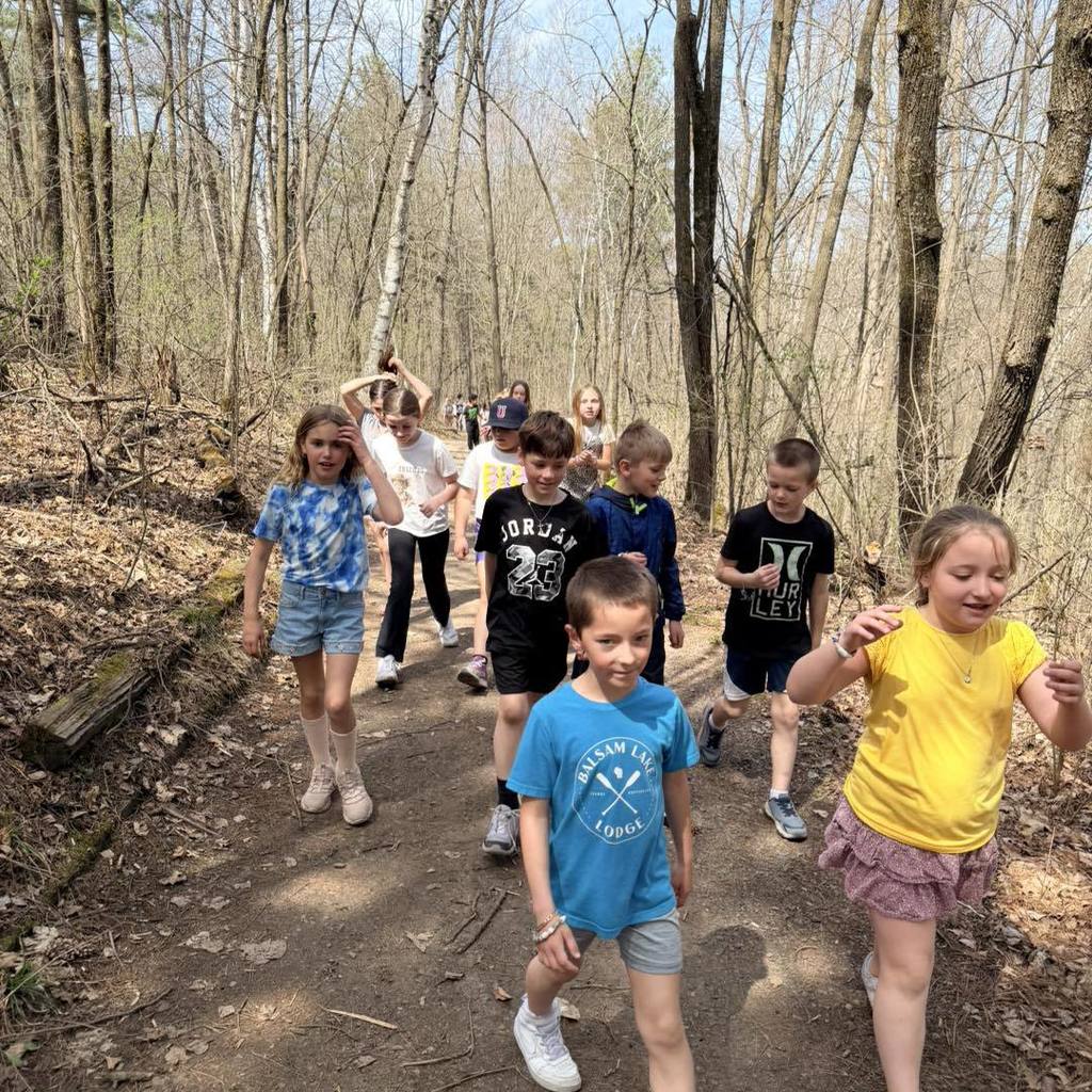 A group of Unity 2nd grade students walking toward the camera on a sunlit dirt nature trail. In the center, a boy in a bright blue shirt leads the way, followed by classmates in yellow and navy shirts, all smiling and enjoying their Earth Day hike through the budding spring woods.