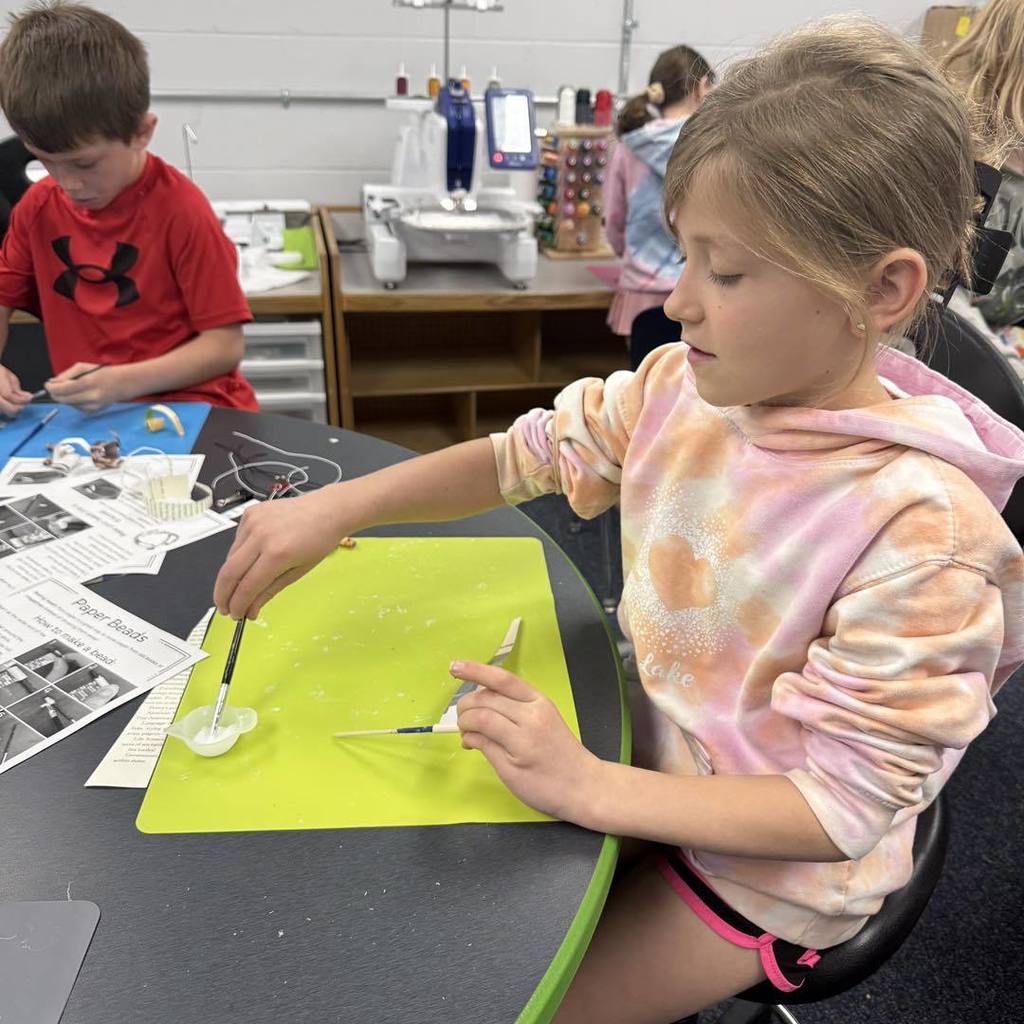 A Unity 2nd grade girl in a tie-dye shirt uses a brush to apply glue to a paper strip on a bright green craft mat. She is carefully crafting a bead from a recycled book page, with instruction sheets and other students working nearby in the classroom.