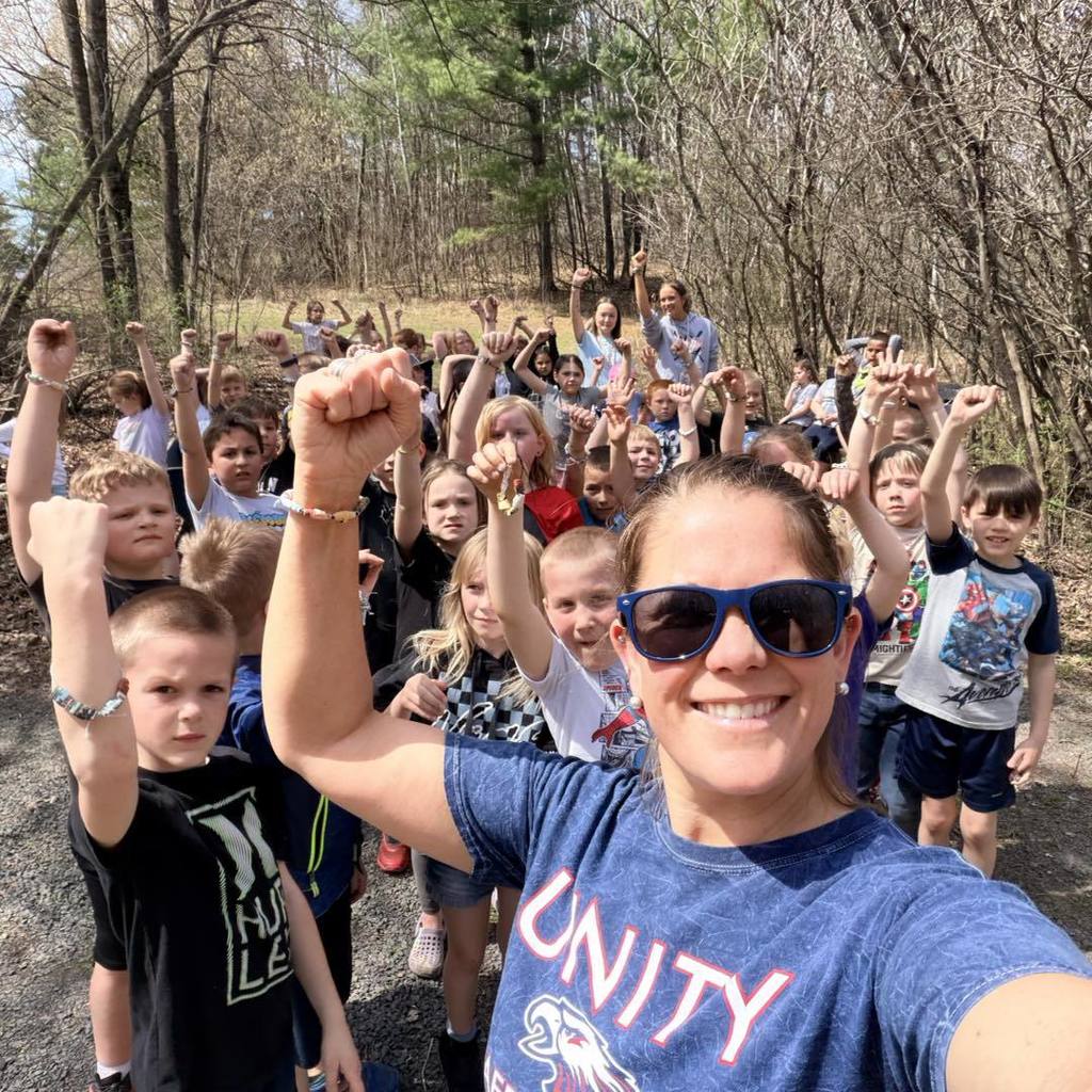 A spirited group selfie taken by a Unity staff member with a large group of 2nd grade students on a wooded nature trail. The staff member, wearing sunglasses and a blue "Unity" t-shirt, smiles alongside students who are cheering with their fists raised in the air to celebrate Earth Day.