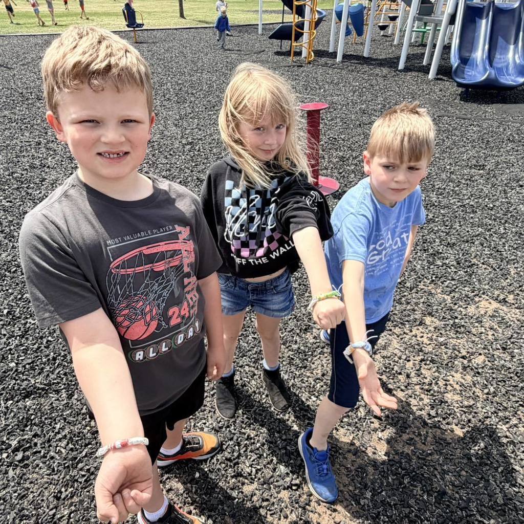 A close-up shot of three Unity 2nd grade students standing on the blacktop and holding their arms out to display handmade bracelets. The bracelets are made from colorful paper beads crafted from recycled book pages, showing a mix of blue, yellow, and patterned designs.
