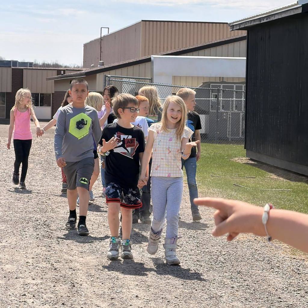 A group of Unity 2nd grade students walking together on a gravel path during their Earth Day celebration. In the foreground, a girl in a white shirt and light blue pants smiles as she walks toward the camera. A hand in the foreground points toward the group, showcasing a handmade paper-bead bracelet.