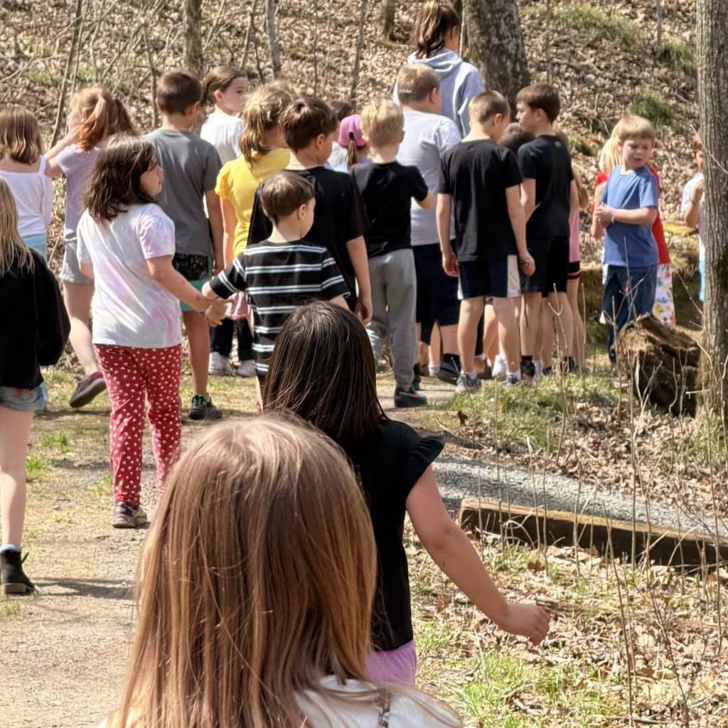 A group of Unity 2nd grade students walking along a wooded nature trail. The photo is taken from behind the group, showing a girl with long brown hair in the foreground following her classmates as they hike through the trees on a sunny Earth Day.