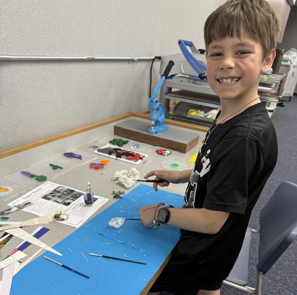 A Unity 2nd grade boy in a black t-shirt smiles for the camera while working on his Earth Day project. He is standing at a table covered with blue craft mats, paper-bead instructions, and various colorful recycled paper strips. In the background, classroom equipment like a heat press and blue tools are visible.