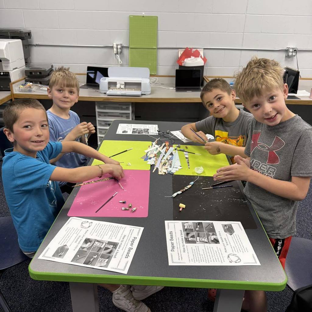 Four Unity 2nd grade boys smile while working together at a classroom craft table. They are using wooden dowels to roll colorful recycled paper strips into beads. The table is organized with bright pink and green craft mats and printed instruction sheets for their Earth Day project.
