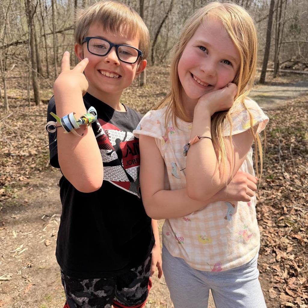 Two Unity 2nd grade students, a boy in a black t-shirt and glasses and a girl in a floral flutter-sleeve shirt, smile while posing on a wooded nature trail. The boy proudly holds up his arm to show off a chunky handmade bracelet made from recycled paper beads, while the girl rests her chin on her hands, also wearing her custom bracelet.