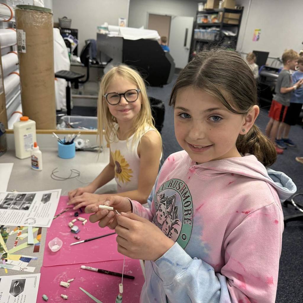 Two Unity 2nd grade girls smile while sitting at a pink craft mat in their classroom. The girl in the foreground, wearing a pink Rushmore hoodie, holds a string of recycled paper beads, while her classmate with glasses works alongside her. They are surrounded by colorful paper strips and instructional guides for their Earth Day project.