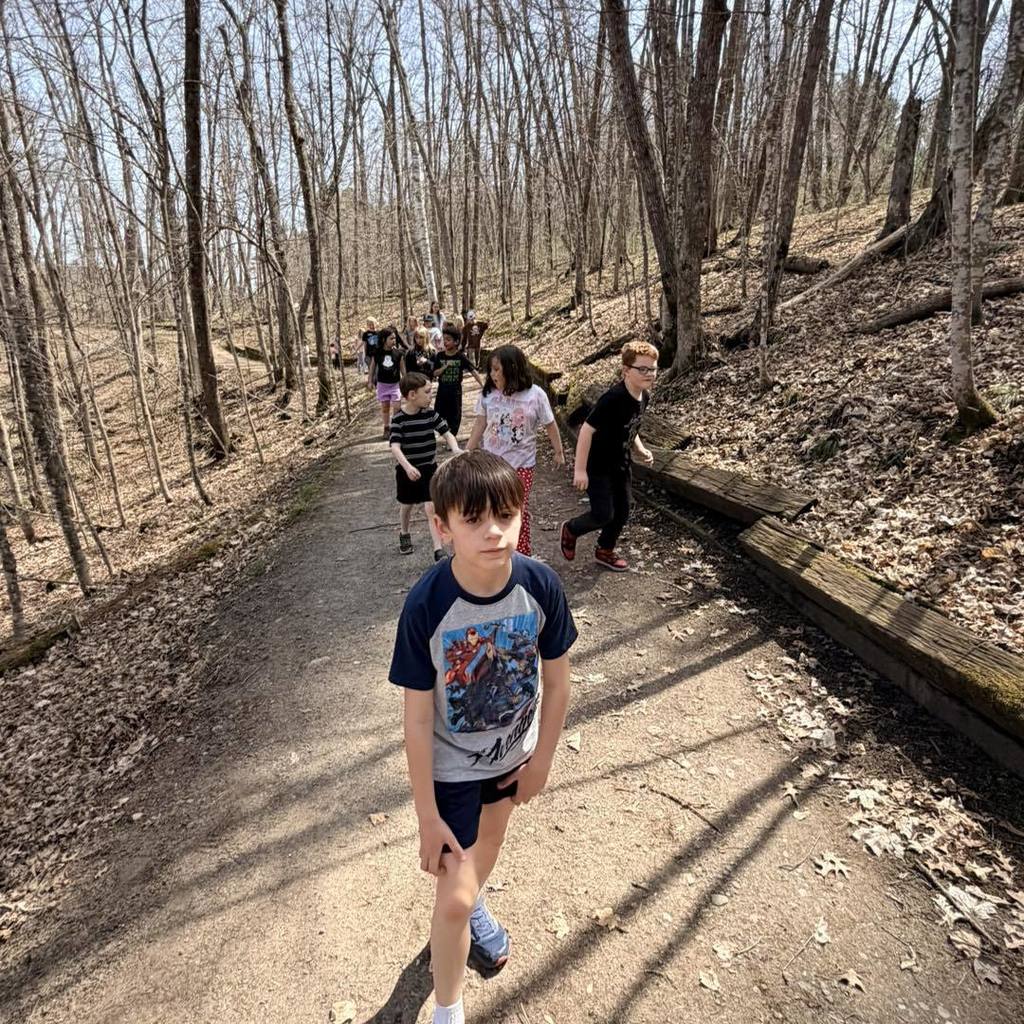 A group of Unity 2nd grade students hiking down a wide, wooded nature trail. A boy in a blue and navy shirt leads the group, followed by several classmates walking single-file. The trail is surrounded by tall, leafless trees and scattered leaves, capturing a bright spring day outdoors.