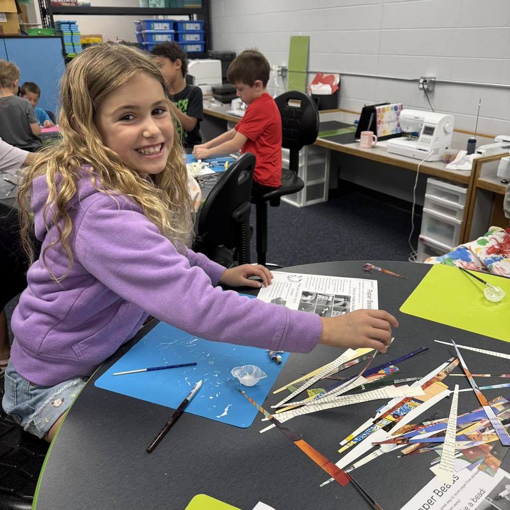A Unity 2nd grade girl with long blonde hair, wearing a purple sweatshirt, smiles brightly while sitting at a craft table. She is making beads from recycled book pages, with colorful paper strips, a blue craft mat, and a small container of glue spread out in front of her.