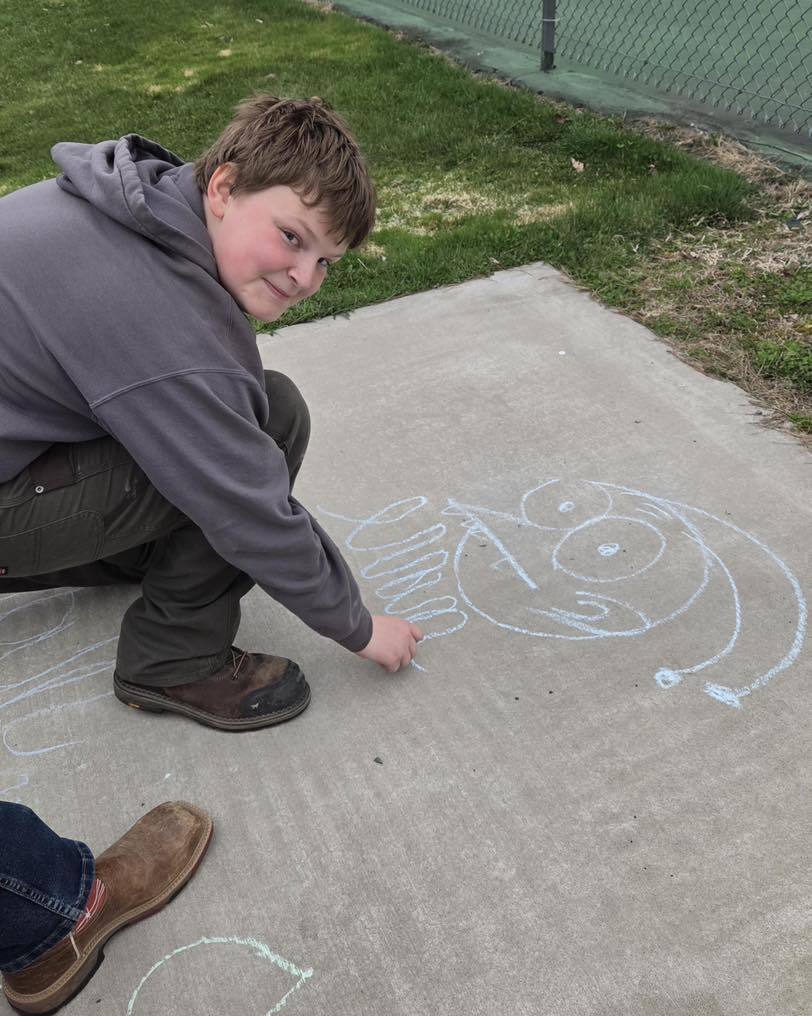A Unity Eagle Time student in a grey hoodie kneels on the sidewalk, smiling at the camera while practicing cursive with chalk. Next to him, he has drawn a large, whimsical face in light blue chalk, showcasing a creative break during Mrs. Schleusner’s handwriting rotation.