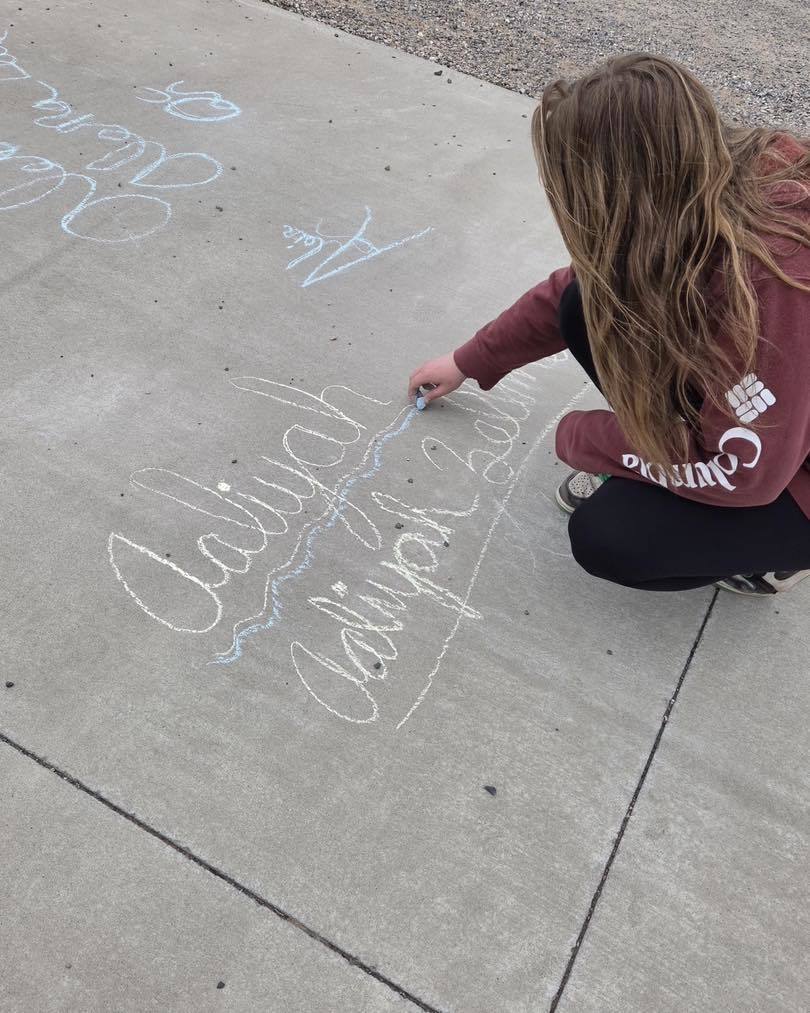 An Eagle Time student at Unity School District practicing her cursive handwriting outdoors using sidewalk chalk. The photo captures her focused on writing her name in large, flowing script during Mrs. Schleusner’s class rotation.