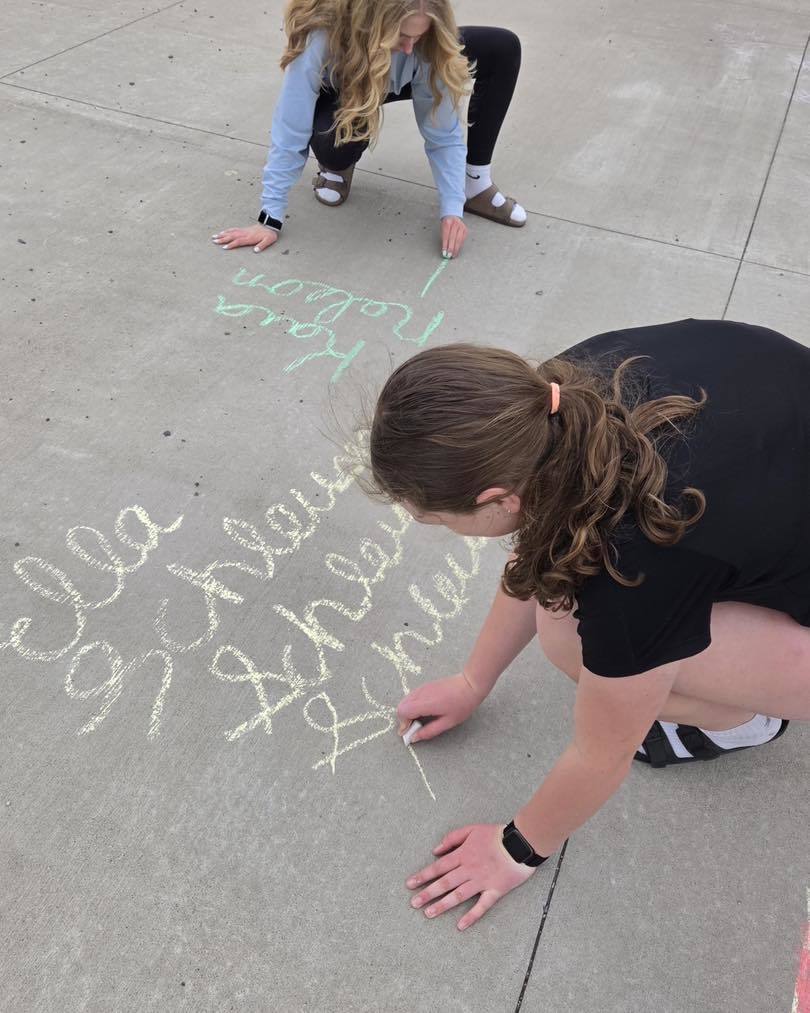 Two Eagle Time students practicing their cursive handwriting skills outdoors. The photo shows the students using colorful sidewalk chalk to write names in elegant script on the concrete.