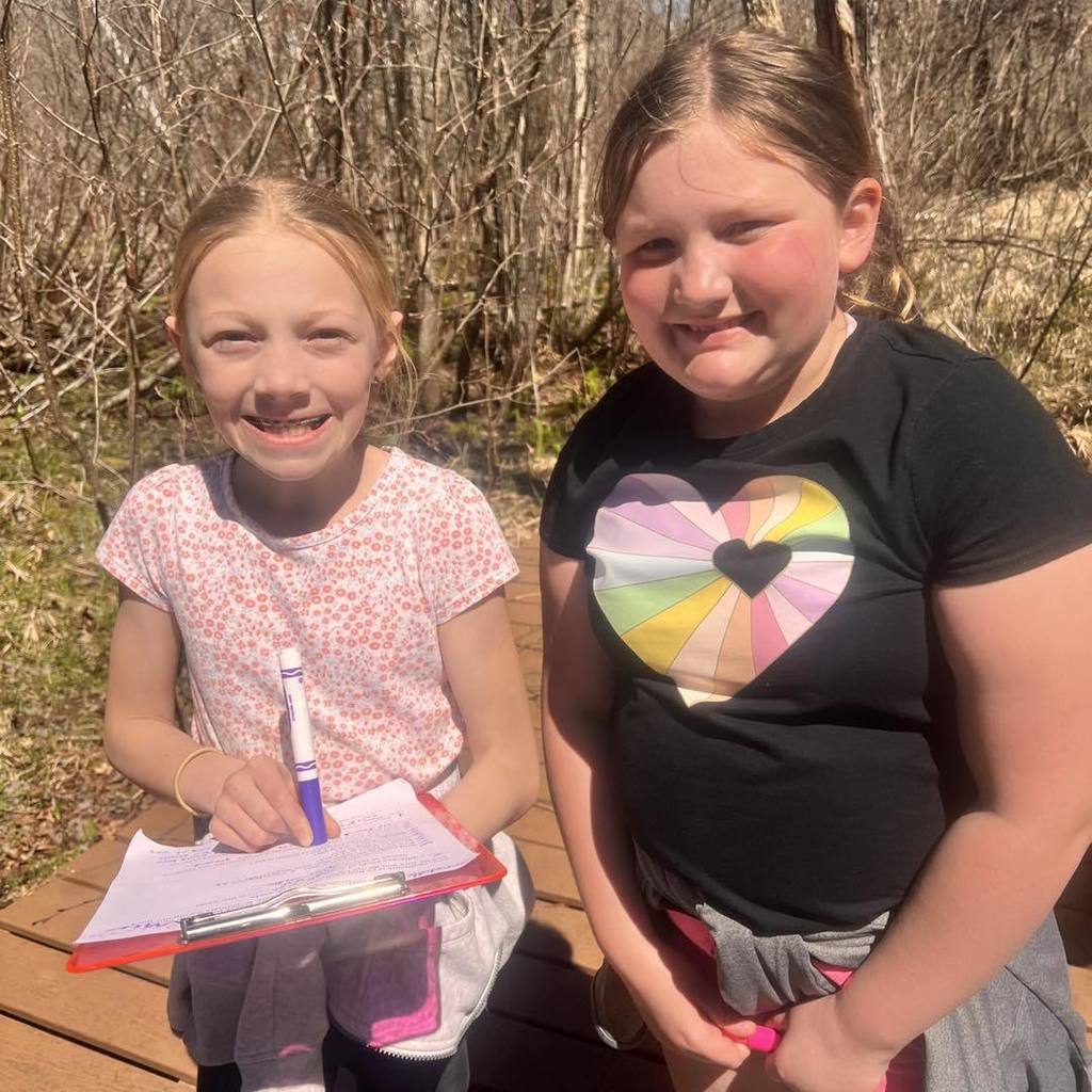 Two smiling elementary students posing on a wooden bridge along a nature trail. One student holds a red clipboard and a blue marker, ready for their outdoor scavenger hunt.