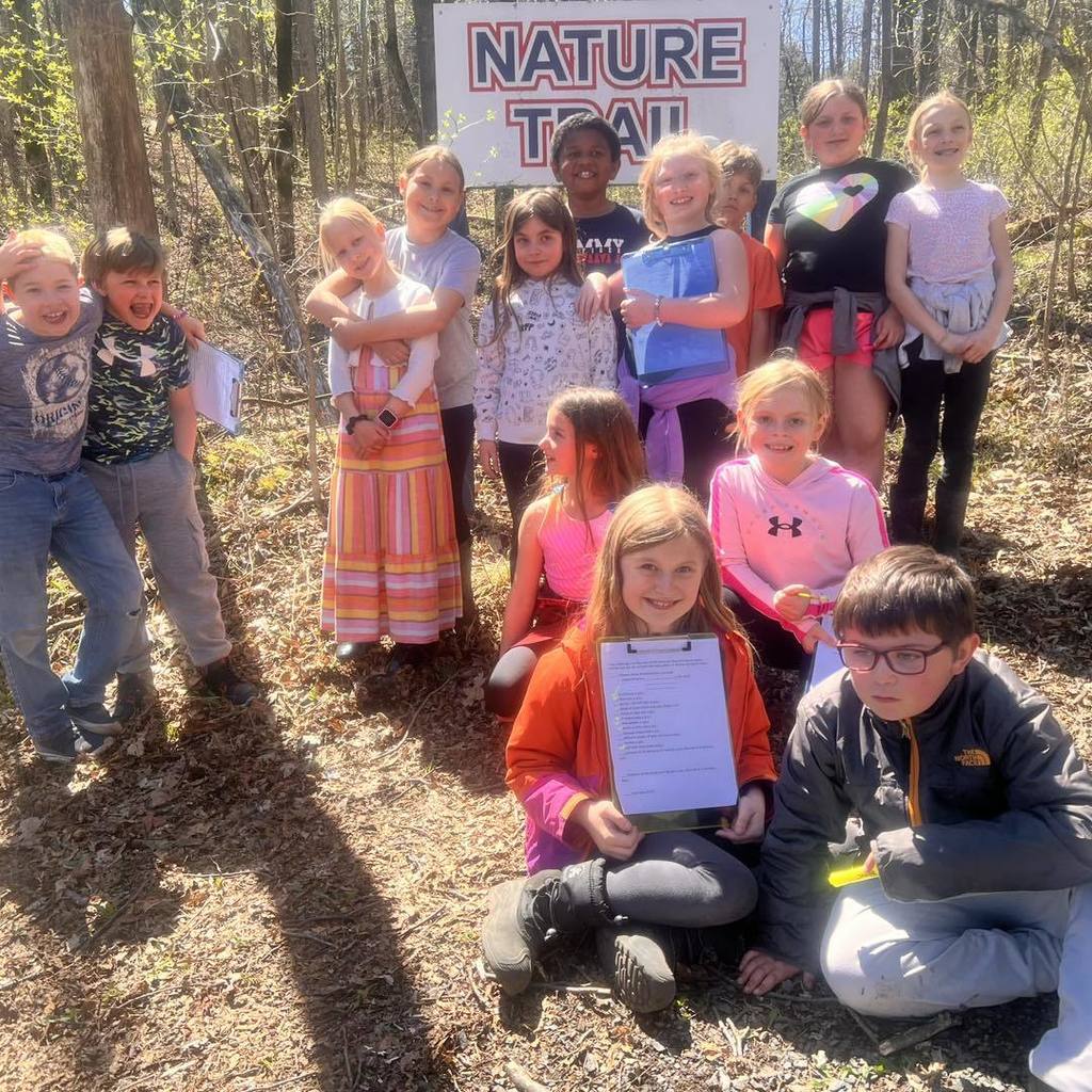 A group of about 15 smiling elementary students posing together on a wooded path. In the background, a wooden sign reads "Nature Trail," and one student in the foreground holds a clipboard with a scavenger hunt list.