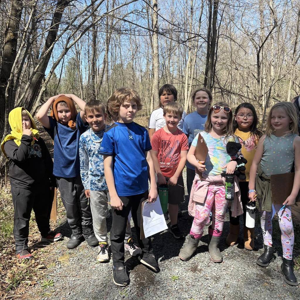 A group of about ten smiling elementary students standing together on a wooded nature trail. Some students are holding clipboards and pencils for their scavenger hunt, and the background shows tall, leafless trees under a clear blue sky.