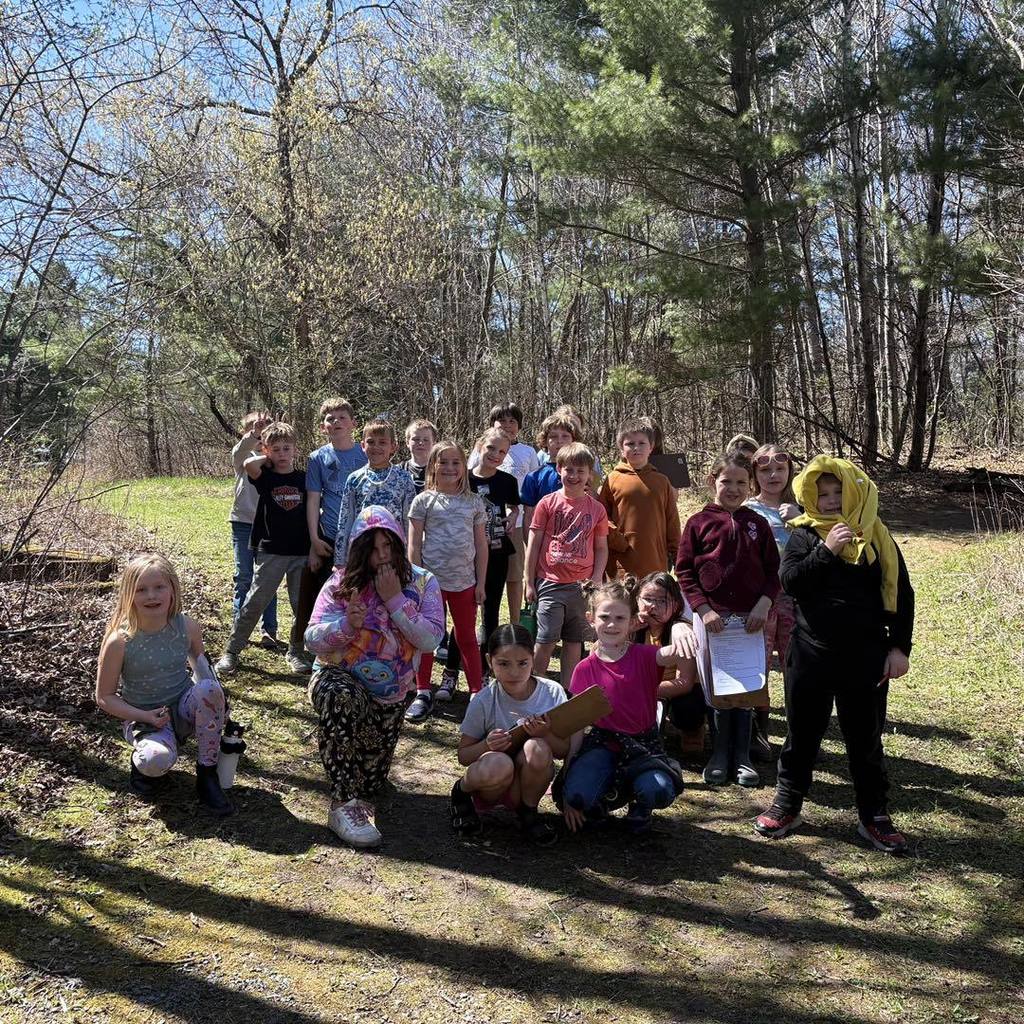 A group of about 20 smiling elementary students posing together in a sunny, wooded area. Several students are holding clipboards and pens for their scavenger hunt, surrounded by tall pine and deciduous trees.