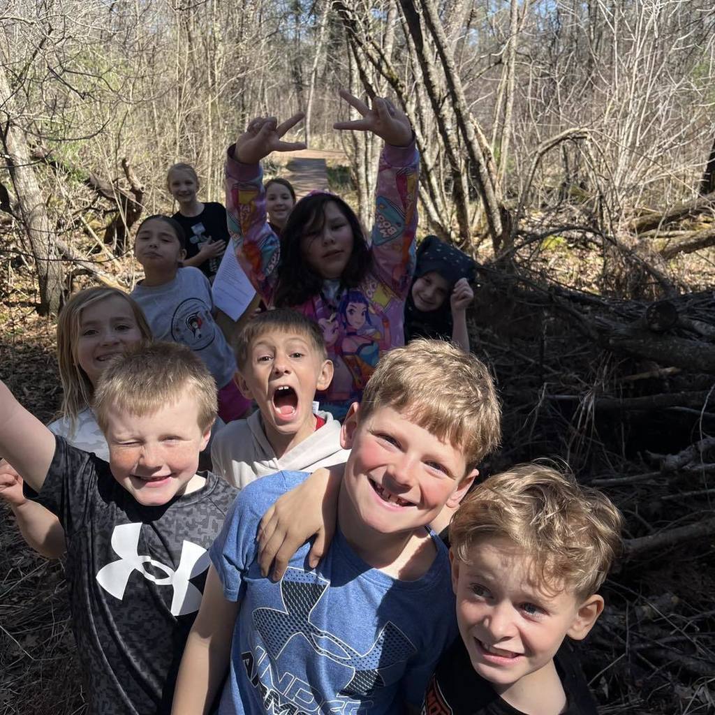 A close-up, joyful group photo of about eight elementary students smiling and making silly faces for the camera. They are huddled together outdoors with a background of thick woods and fallen branches.