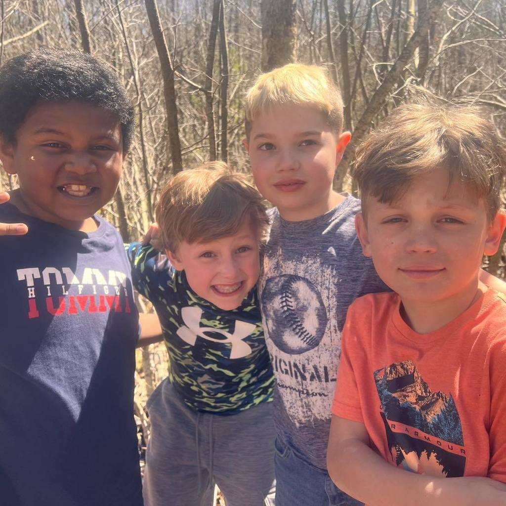 A close-up photo of four young boys smiling and huddling together for a group shot on the nature trail. They are dressed in casual t-shirts, and the background shows the bright, sun-dappled woods.