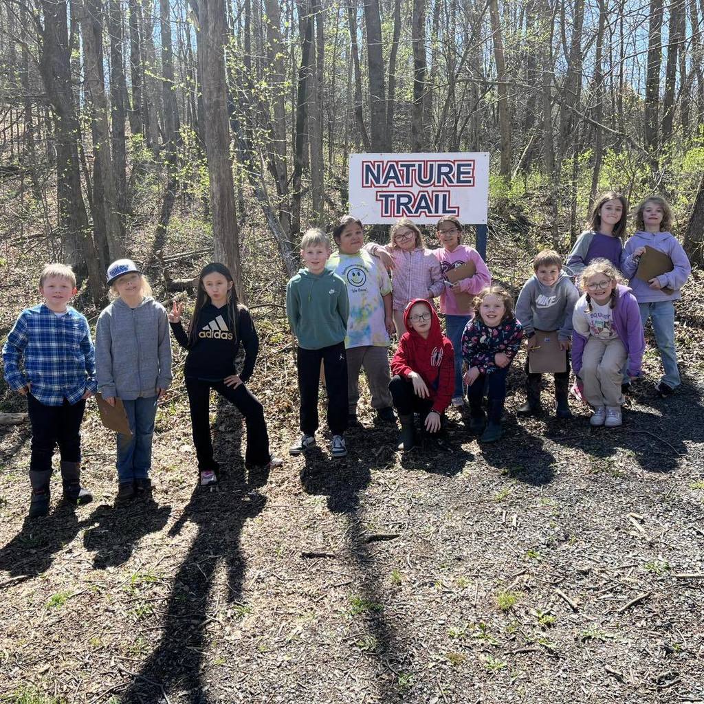A group of elementary students gathered around a wooden sign that says "Nature Trail" at the start of a wooded path. Some students are holding clipboards, and everyone is smiling for the camera.