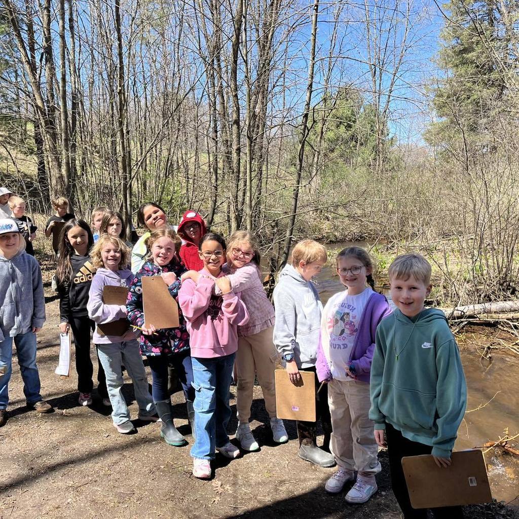 A group of about ten elementary students standing on a dirt path next to a small stream. Two girls in the center are hugging and smiling, while other students hold clipboards and pencils, prepared for their outdoor scavenger hunt.