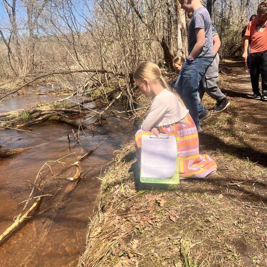 A young student wearing a colorful striped dress kneels at the edge of a small stream. She is holding a green clipboard and looking down at the water, recording observations for the scavenger hunt.
