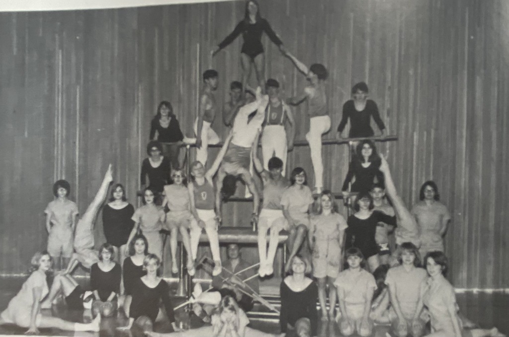 A vintage black-and-white group photo of a gymnastics team posing in a gymnasium. The students are arranged in a large pyramid formation using wooden benches and bars, with some athletes performing handstands and poses while others sit or stand in rows.
