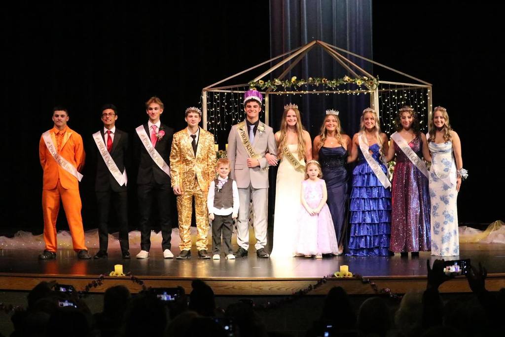 The 2026 Unity School District Prom Court posing on stage under a wooden arch decorated with greenery and fairy lights. The group includes students in formal gowns and suits with "Prom Court," "Prom King," and "Prom Queen" sashes, flanked by two young junior royalty members.
