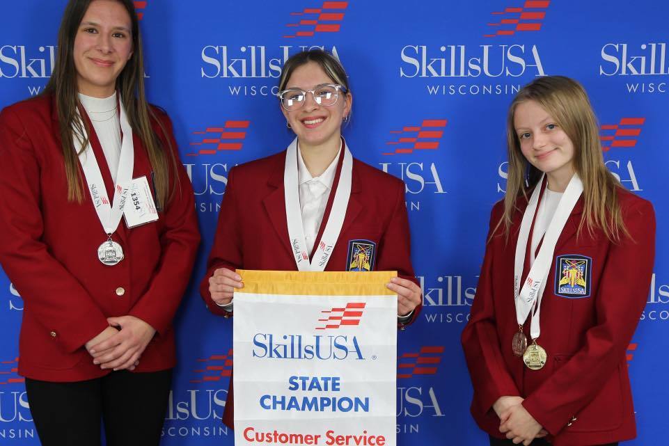 Three Unity students in red SkillsUSA blazers posing in front of a blue SkillsUSA Wisconsin backdrop. The student in the center holds a "State Champion" banner for Customer Service, and all three are wearing competition medals.