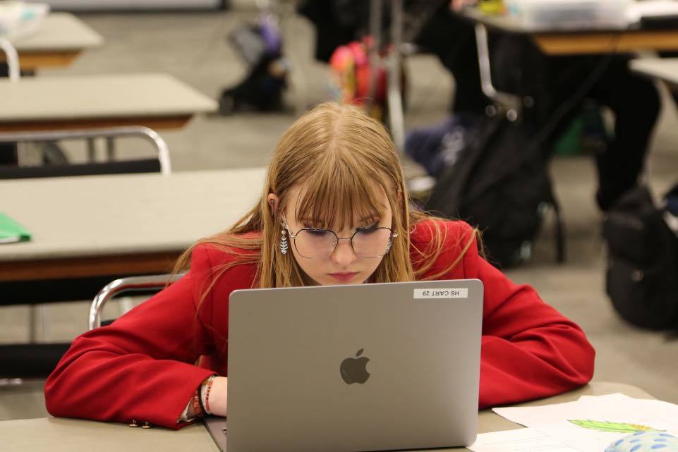 A Unity student wearing a red SkillsUSA blazer and glasses, sitting at a table and working intently on a laptop during the state competition in Madison.