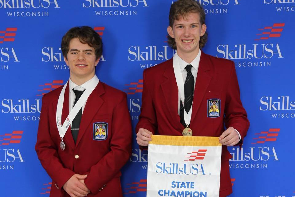 Two Unity students in red SkillsUSA blazers posing in front of a blue SkillsUSA Wisconsin backdrop. The student on the right is smiling and holding a white and blue "SkillsUSA State Champion" banner, and both students are wearing competition medals.
