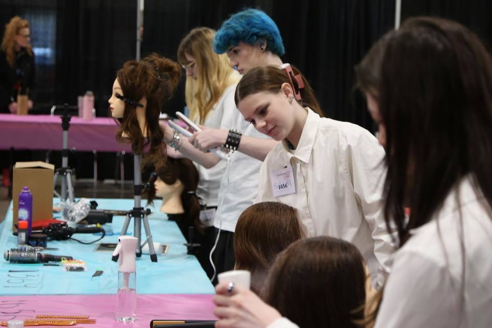 A group of SkillsUSA students in white shirts working at a long table during a competition. In the foreground, a student uses a flat iron on a mannequin head's hair, while other competitors and mannequins are visible in the background of the busy event space.