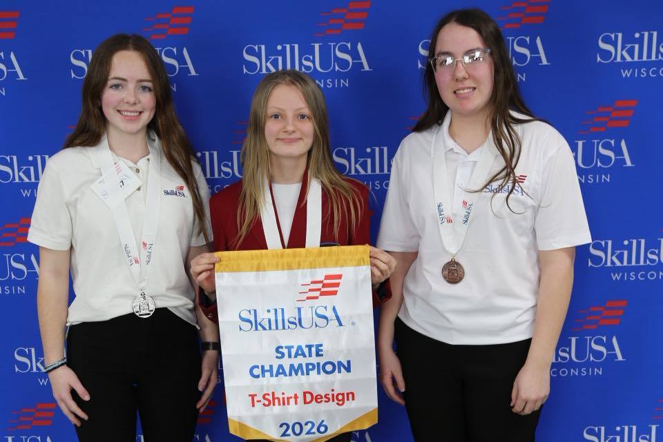 Three Unity students standing in front of a blue SkillsUSA Wisconsin backdrop. The student in the center is wearing a red blazer and holding a "State Champion T-Shirt Design 2026" banner, while the two students flanking her wear white polo shirts and competition medals.