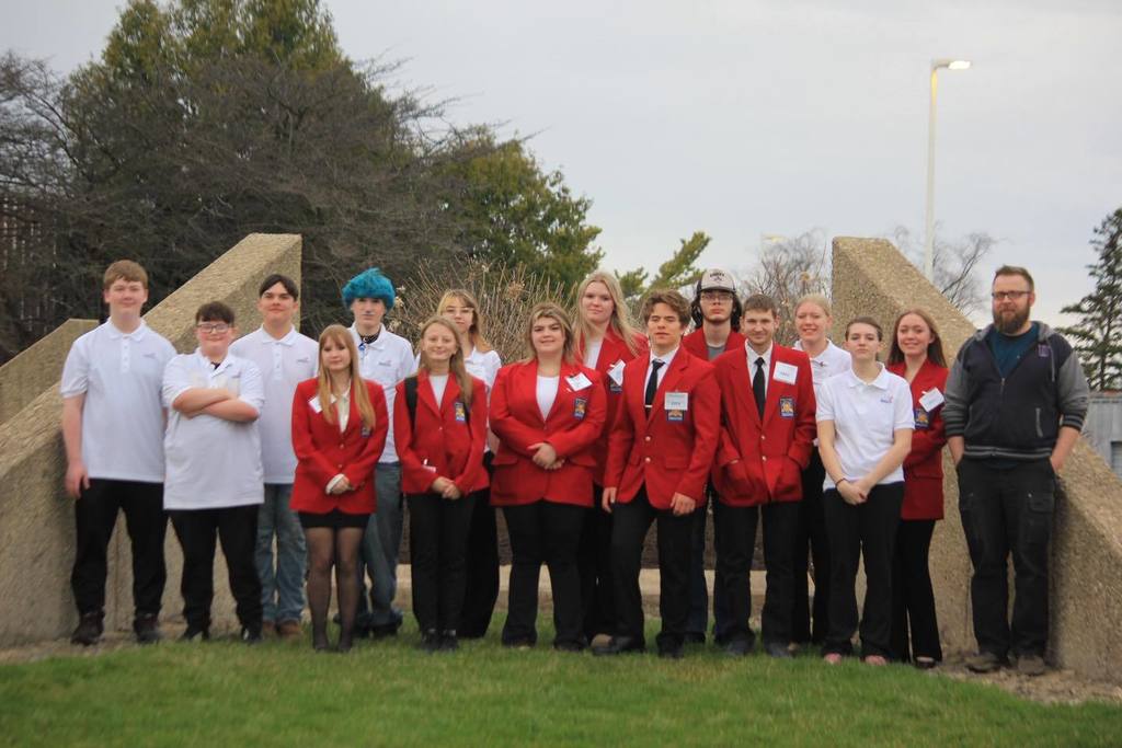A group photo of fifteen Unity SkillsUSA students and their advisor standing outside in front of a stone monument. Several students are wearing red SkillsUSA blazers, while others are in white and grey polo shirts.