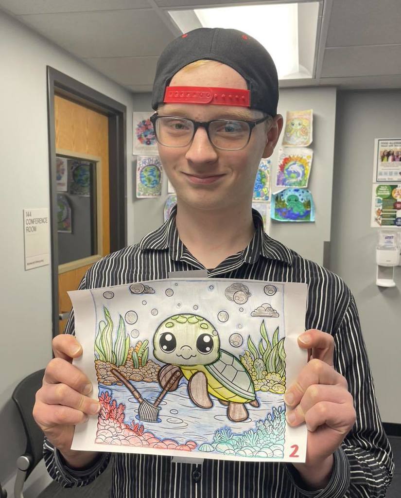 A student wearing a striped button-down shirt and a black baseball cap with a red brim, smiling while holding his Earth Day coloring entry. The artwork features a cartoon sea turtle swimming near the ocean floor with bubbles and seaweed. He is standing in an office or classroom with other entries displayed on the wall behind him.