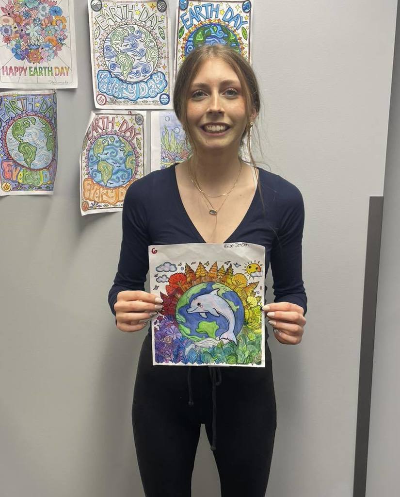 A student in a dark long-sleeve shirt smiling and holding her Earth Day coloring entry, which features a dolphin jumping in front of a colorful globe surrounded by flowers. She is standing in front of a wall showcasing other student art submissions.