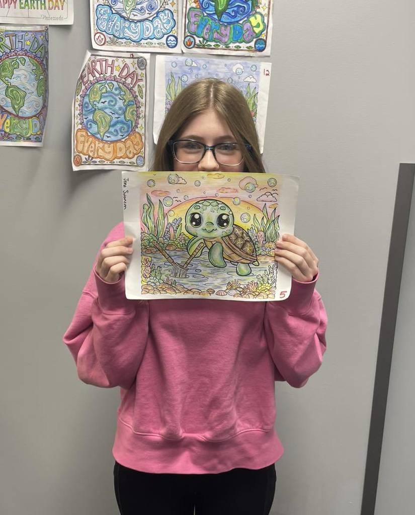 A student in a pink sweatshirt holding up his Earth Day coloring entry, which features a detailed drawing of a sea turtle swimming near the ocean floor. He is standing in front of a wall where other Earth Day coloring contest entries are displayed.