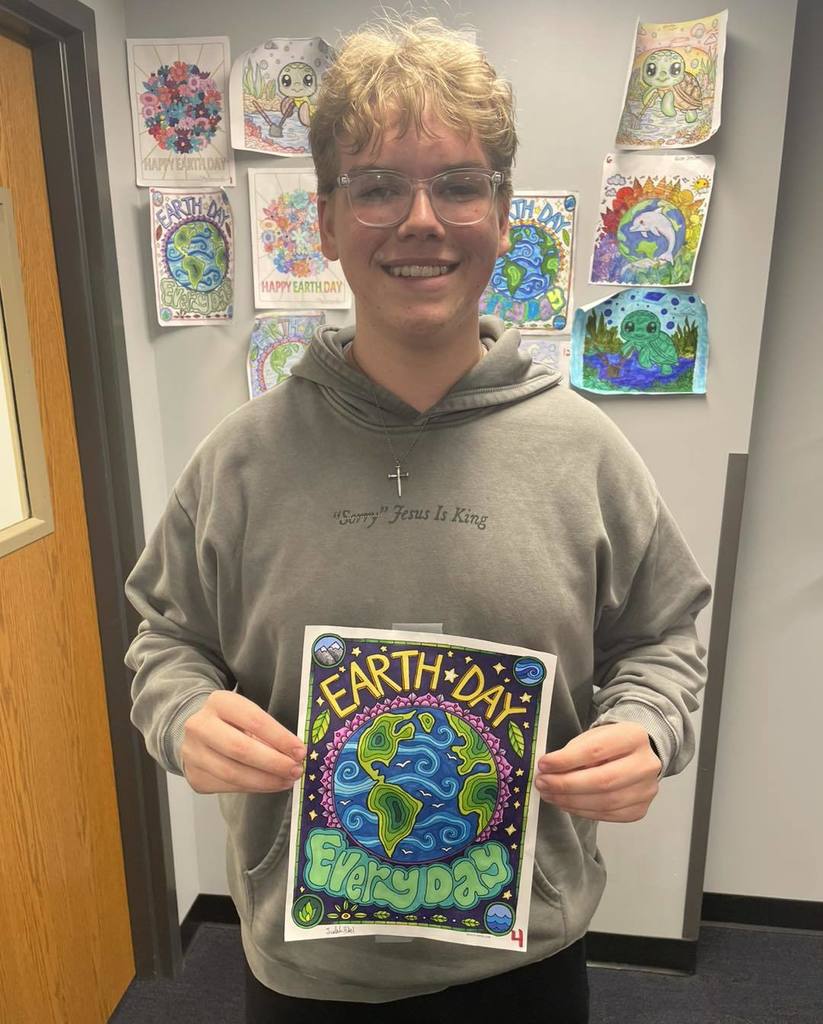 A high school student wearing a grey hoodie and glasses, smiling and holding up his winning Earth Day coloring page. The artwork is vibrant with blue and green swirls and the words "Earth Day Everyday." Other colorful entries are displayed on the wall behind him.