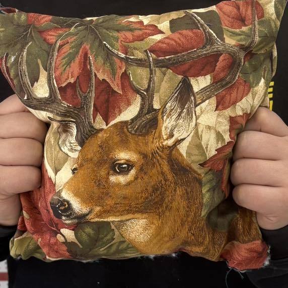 A close-up of a small handmade pillow or fabric square being held by a student. The fabric features a detailed, realistic print of a buck's head with large antlers, surrounded by a pattern of red and orange autumn maple leaves.