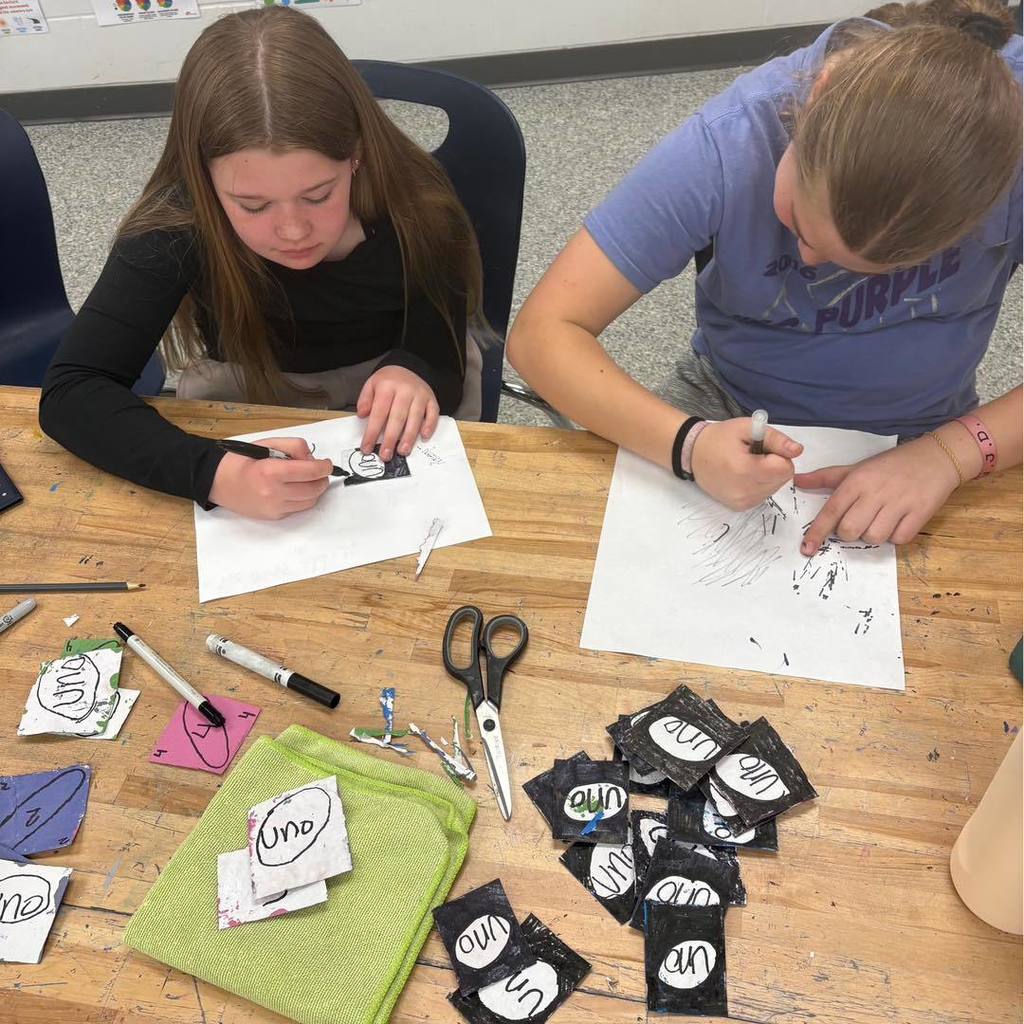 Two students working at a wooden table on a textiles project. They are using markers to draw on white paper, surrounded by small fabric scraps featuring "Uno" card designs and a pair of scissors.