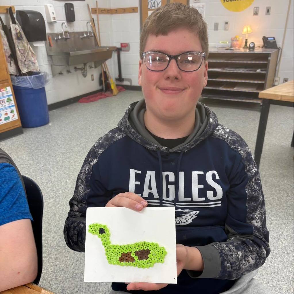 A student in a navy blue "EAGLES" hoodie and glasses, smiling while holding up a square white canvas featuring a green and brown latch-hook or textile design in the shape of a dinosaur or creature. He is sitting in a classroom with a sink and cabinets in the background.