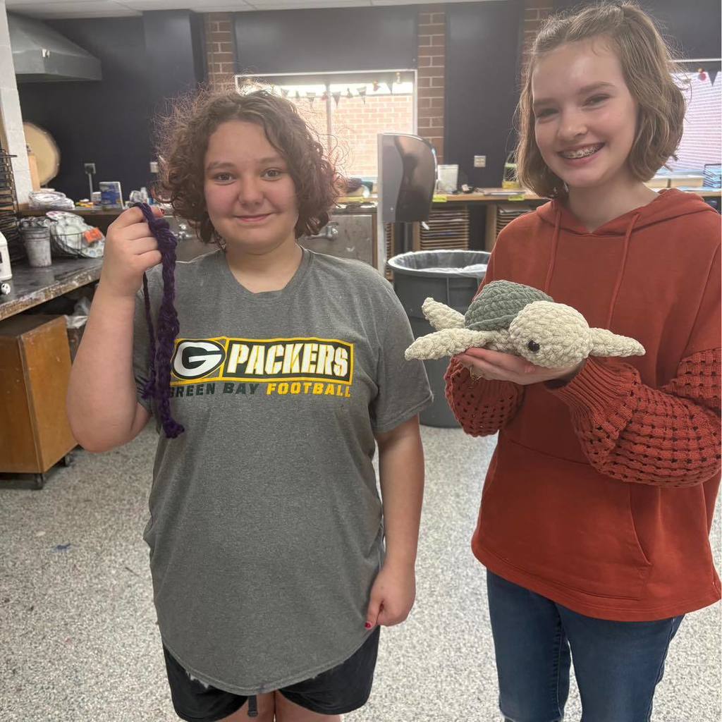 Two Unity students standing in a classroom and smiling while showing off their textile projects. The student on the left, wearing a grey Green Bay Packers t-shirt, holds up a purple crocheted piece, while the student on the right, in a red hoodie, holds a small crocheted sea turtle.