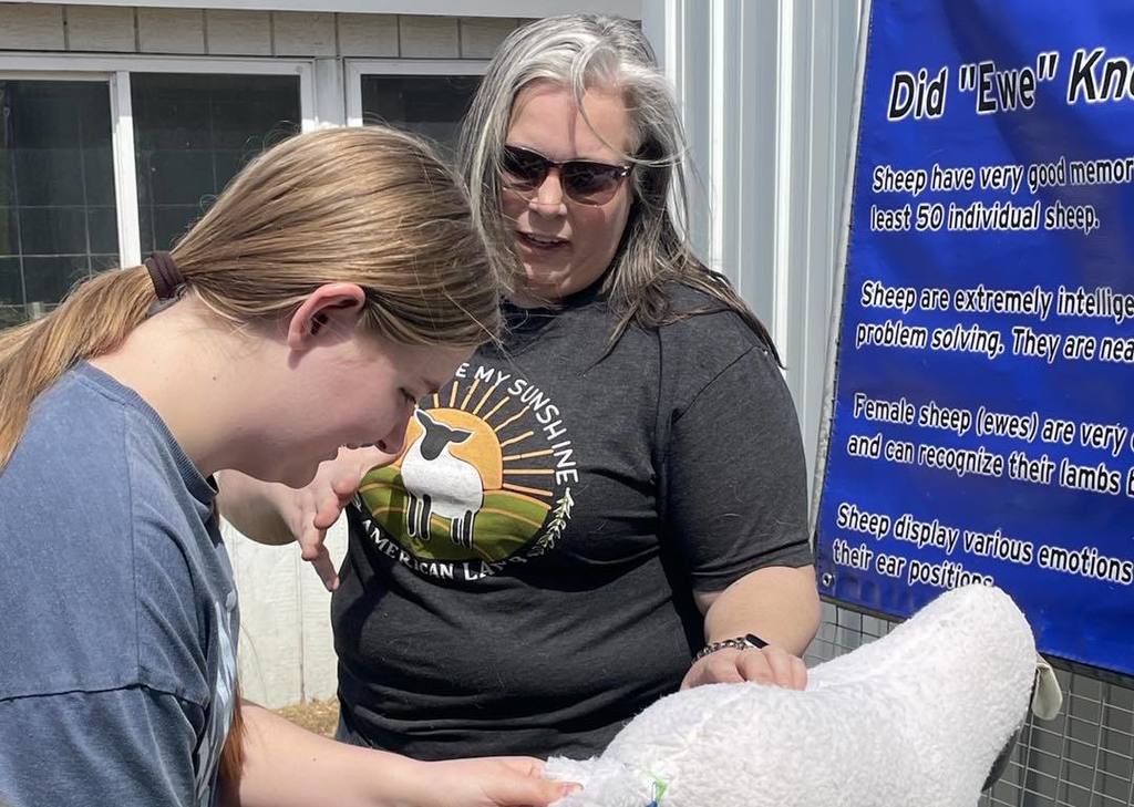 An instructor in a black t-shirt and sunglasses assists a student in a blue t-shirt as they examine a fluffy white sheep during a hands-on learning station. In the background, an educational sign titled "Did 'Ewe' Know" provides facts about sheep intelligence and memory.