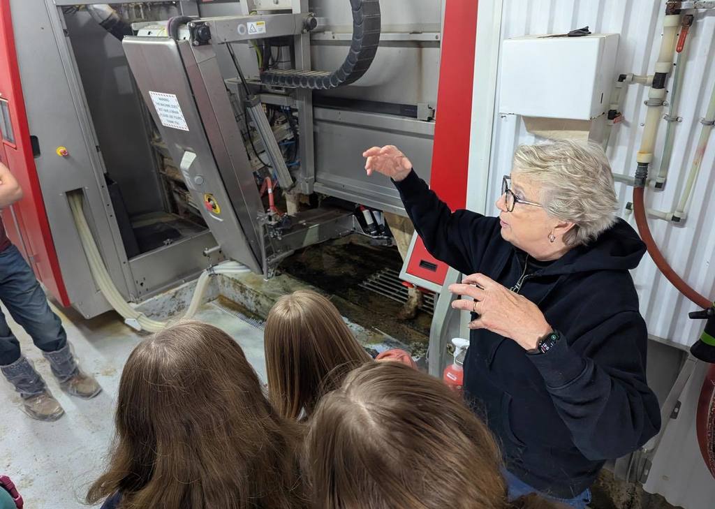 An instructor in a black hoodie and glasses gestures toward a piece of modern dairy machinery while explaining its function to three students. The students are listening intently during a tour of the state-of-the-art facility at Four Cub Farms.
